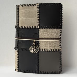 Puede incluir: Una cubierta de diario rectangular con un diseño de patchwork de paneles de cuero negro, gris y beige con costuras oscuras visibles. Una fina banda elástica blanca envuelve el centro, sosteniendo un dije de metal dorado con un diseño calado.