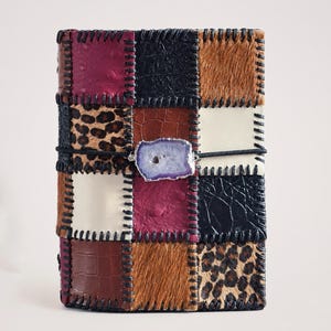 Peut inclure: Une couverture de journal rectangulaire présentant un design en patchwork de diverses textures, incluant un imprimé léopard, du similicuir marron et noir, et des panneaux bordeaux texturés. Les pièces sont assemblées par des coutures noires.