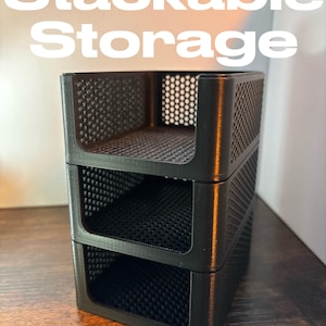 May include: Black stackable storage containers with a perforated design. The containers are stacked on top of each other. The text "Stackable Storage" is in white above the containers.