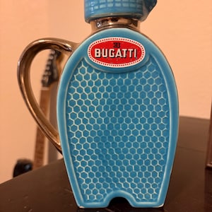 May include: A blue ceramic pitcher with a gold handle and rim. The pitcher features a honeycomb pattern and a red and white Bugatti logo. The pitcher is a decorative item.