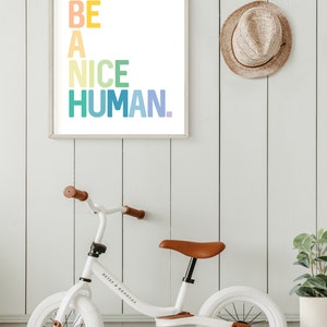 May include: A white framed print with the text "BE A NICE HUMAN." in rainbow colors, a white balance bike with brown leather seat and a brown straw hat hanging on a white wood wall.