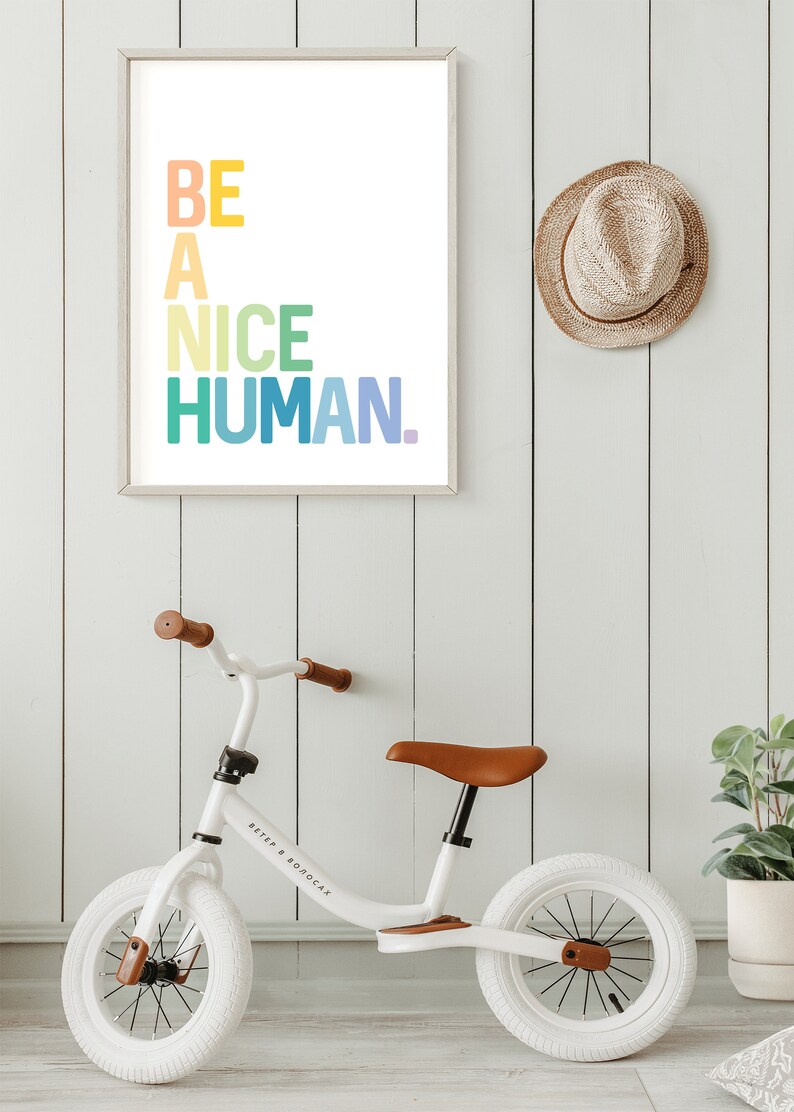 May include: A white framed print with the text "BE A NICE HUMAN." in rainbow colors, a white balance bike with brown leather seat and a brown straw hat hanging on a white wood wall.