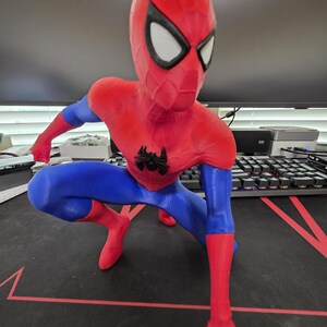 May include: A red, blue, and black Spider-Man figurine in a crouching pose. The figurine has a red head and torso, blue arms and legs, and black accents. The Spider-Man logo is on the chest. The eyes are white.