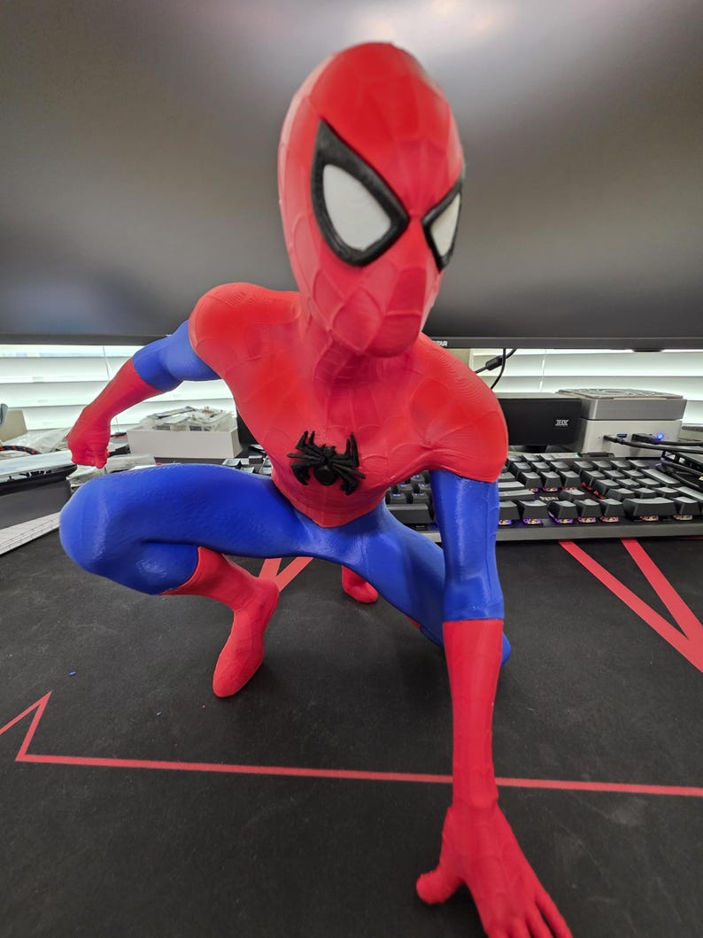 May include: A red, blue, and black Spider-Man figurine in a crouching pose. The figurine has a red head and torso, blue arms and legs, and black accents. The Spider-Man logo is on the chest. The eyes are white.