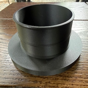 Puede incluir: Un adaptador cilíndrico negro mate con una base circular ancha y una sección de tubo superior más estrecha. El objeto tiene un acabado liso y uniforme con una costura horizontal distintiva donde se unen los dos componentes circulares.