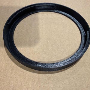 Puede incluir: Un anillo circular negro con una superficie texturizada y un borde interior elevado. El objeto presenta un centro hueco y una base plana, mostrando un acabado negro mate consistente en toda su estructura. El diseño incluye pequeñas pestañas.