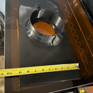 Puede incluir: Un adaptador de escape rectangular negro impreso en 3D con un puerto circular y dos perillas pequeñas, con el texto 'comgrow3Dprinting.com' en el lateral. Una cinta métrica amarilla se coloca sobre la base, mostrando un ancho de unos 26,7 cm.