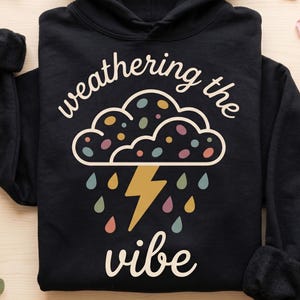 May include: Black hoodie with the phrase "weathering the vibe" in white script. The design features a cloud with colourful dots, raindrops, and a yellow lightning bolt. The hoodie has a hood and long sleeves.