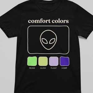 Könnte beinhalten: Schwarzes T-Shirt mit dem Text "comfort colors" über einem weißen Alien-Kopf-Design. Darunter befinden sich vier Farbfelder mit den Bezeichnungen "beam", "glow", "float" und "home".