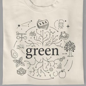 以下が含まれることがあります： クリーム色のTシャツ。黒いグラフィックデザインで、「green」の文字が、木、アボカド、お金のシンボルなど、さまざまなイラストで囲まれています。Tシャツはきれいに折りたたまれています。