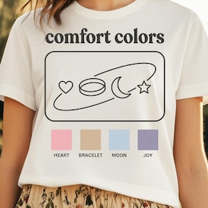 May include: White t-shirt with the words "comfort colors" above a graphic of a heart, ring, crescent moon, and star. Below the graphic are four color swatches labeled "Heart," "Bracelet," "Moon," and "Joy."