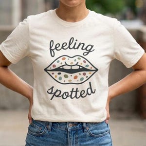 May include: A cream-coloured t-shirt with the words "feeling spotted" in black script. The shirt features a graphic of lips with colourful spots. The model is wearing blue jeans.