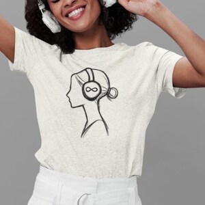 May include: A light grey t-shirt with a black line drawing of a woman's profile wearing headphones with an infinity symbol on the earcups. The design is centered on the shirt.