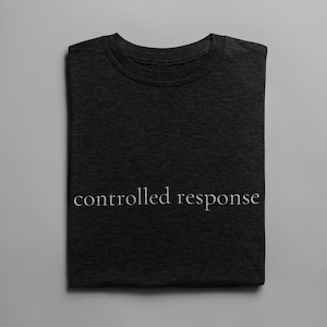 May include: Dark gray t-shirt with the words "controlled response" printed in a light color across the front. The shirt is folded neatly and centered on a gray background.