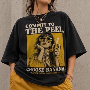 Könnte beinhalten: Ein schwarzes Kurzarm-T-Shirt mit einer gelben rechteckigen Grafik, die eine Person mit Sonnenbrille zeigt, die eine Banane hält. Der Text lautet COMMIT TO THE PEEL, CHOOSE BANANA und NATURE'S MOST UNHINGED SNACK in Vintage-Schrift.