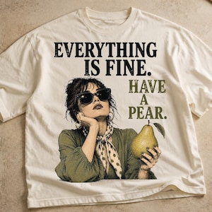 May include: An off-white t-shirt featuring a graphic of a person wearing sunglasses and a polka-dot neck scarf, holding a pear. The text reads "EVERYTHING IS FINE. HAVE A PEAR." The design is printed in black and olive green ink on the front of the shirt.