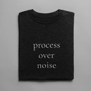 May include: Dark gray t-shirt with the words "process over noise" printed in white. The shirt is folded neatly and has a crew neck. The text is centered on the front of the shirt.