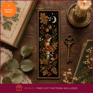 May include: A rectangular cross-stitch bookmark featuring a black background with a central brown mouse sitting on a red and white spotted mushroom, surrounded by autumn oak leaves, acorns, small red berries, and a white crescent moon.