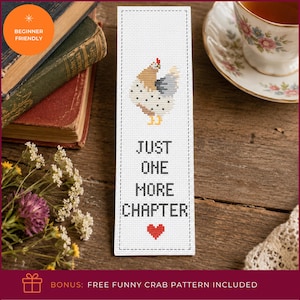 May include: A rectangular white fabric bookmark featuring a cross-stitched design of a speckled chicken in shades of brown, grey, and yellow. Below the chicken, the text reads JUST ONE MORE CHAPTER in black, followed by a small red heart.