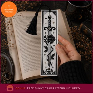 Op de afbeelding: Een rechthoekige kruissteek boekenlegger met een zwart-wit ontwerp van een zwaard verstrengeld met een roos, geaccentueerd met kleine gouden sterretjes. De boekenlegger is afgewerkt met een zwarte kwast aan de bovenkant en een decoratieve rand.