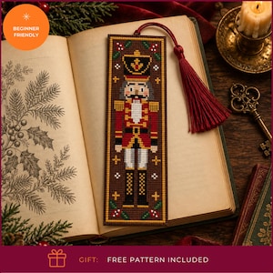 May include: A rectangular cross-stitch bookmark featuring a nutcracker soldier in a red, gold, and black uniform on a brown background. The bookmark is finished with a dark red tassel attached to the top and is displayed resting on an open book.
