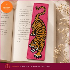 May include: A rectangular needlepoint bookmark featuring a vibrant pink background with a detailed tiger design in shades of orange, black, and white. The tiger is depicted in a pouncing position with its mouth open, showcasing sharp white teeth.