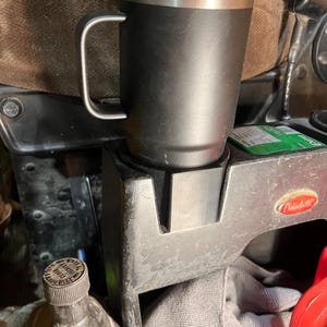 May include: A black insulated travel mug with a silver rim and handle. The mug is cylindrical and sits in a vehicle's cup holder. The mug is designed to keep beverages hot or cold for extended periods.