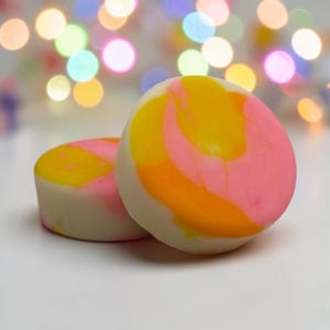 Könnte beinhalten: Zwei runde, weiße Objekte mit bunten Wirbeln aus Gelb, Orange und Rosa. Die Objekte befinden sich auf einer weißen Oberfläche, mit einem verschwommenen Hintergrund aus bunten Bokeh-Lichtern.
