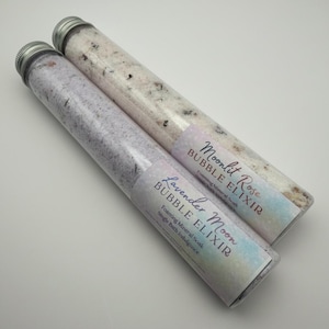 Może przedstawiać: Dwie przezroczyste tuby ze srebrnymi nakrętkami, wypełnione solami do kąpieli. Jedna tuba jest w kolorze lawendy, z etykietą "Lavender Moon Bubble Elixir". Druga jest biała, z etykietą "Moonlit Rose Bubble Elixir". Obie etykiety zawierają "Foaming Mineral Soak".