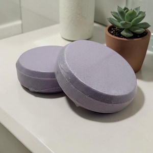 Pode incluir: Duas barras de sabão redondas, cor lavanda, são exibidas em uma superfície branca. Os sabonetes são embalados em plástico transparente. Um pequeno vaso de suculenta e um recipiente de cerâmica branco estão ao fundo.