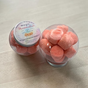 May include: Two clear, spherical containers filled with pink, heart-shaped bath cubes. One container has a silver lid with the text "magic Marshmallows" and "Whipped Body Wash Cubes". The other container is open, showcasing the fluffy cubes.