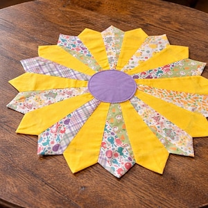 May include: A handmade, circular placemat with a sunflower design. The centre is a solid purple circle, surrounded by yellow fabric petals. The petals alternate between solid yellow and patterned fabrics.