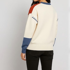 以下が含まれることがあります： クリーム色のセーター、カラーブロックデザイン。セーターは、錆色の肩パネル、青い袖口、青いアクセントが特徴です。袖には装飾的なステッチパターンがあります。セーターは黒いパンツと組み合わせています。