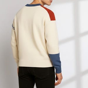 以下が含まれることがあります： クリーム色のセーター、カラーブロックデザイン。赤い肩のアクセント、青い袖口、青いネックラインのトリムが特徴です。セーターはリブ編みの裾と袖口が付いています。モデルは黒いパンツを着用しています。