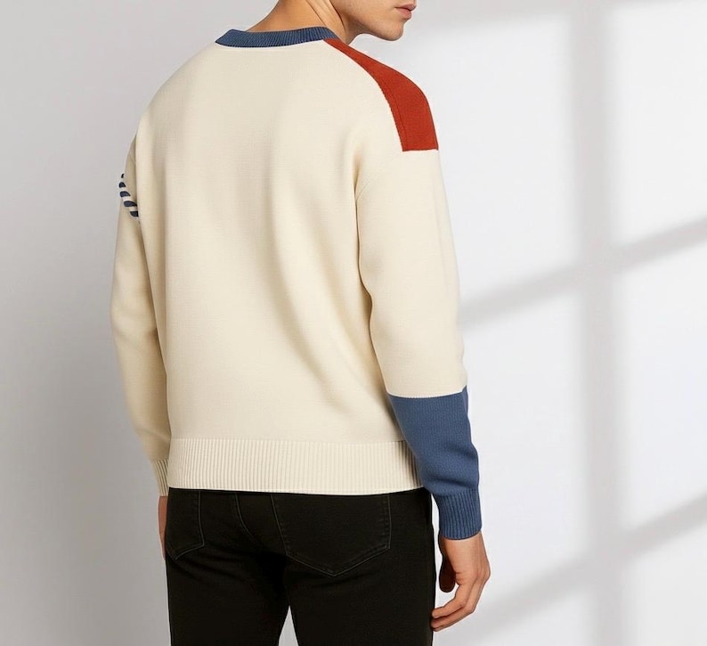 以下が含まれることがあります： クリーム色のセーター、カラーブロックデザイン。赤い肩のアクセント、青い袖口、青いネックラインのトリムが特徴です。セーターはリブ編みの裾と袖口が付いています。モデルは黒いパンツを着用しています。