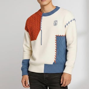 May include: Cream-colored sweater with color block design. Features a rust-colored cable knit panel on the shoulder, blue and cream sections, and blue stitching details. A small emblem is on the chest.