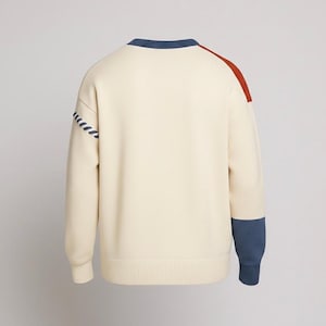 以下が含まれることがあります： クリーム色のセーター。青と錆色のアクセントが特徴。青いネックライン、肩の錆色のストライプ、袖口の青いカフス。左袖には装飾的なステッチデザインがあります。