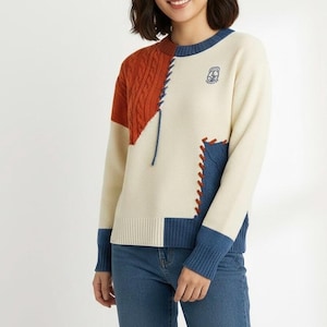 以下が含まれることがあります： クリーム色のセーター、カラーブロックデザイン。 焦げたオレンジ色のケーブルニット部分、デニム風の青いパッチ、首と袖口の青いトリムが特徴です。 胸には小さな刺繍エンブレムが見えます。 セーターは青いジーンズと組み合わせています。