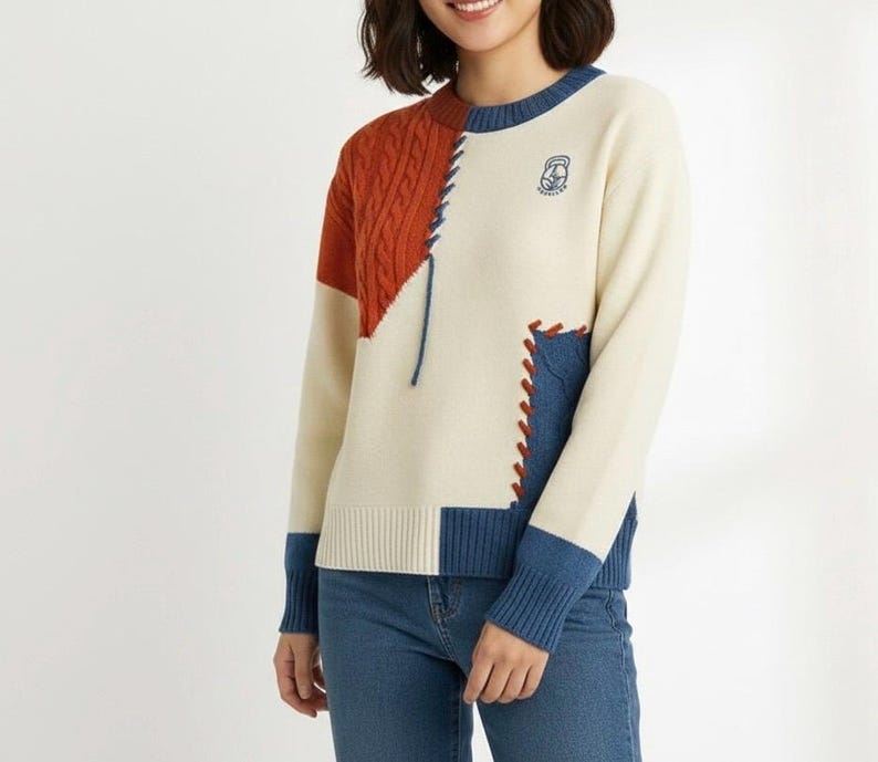 以下が含まれることがあります： クリーム色のセーター、カラーブロックデザイン。 焦げたオレンジ色のケーブルニット部分、デニム風の青いパッチ、首と袖口の青いトリムが特徴です。 胸には小さな刺繍エンブレムが見えます。 セーターは青いジーンズと組み合わせています。