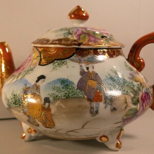 Pode incluir: Bule decorativo de porcelana com tampa, apresentando uma cena pintada à mão de figuras em trajes tradicionais. O bule tem uma base branca com detalhes em laranja na alça, no bico e na tampa. O bule tem aproximadamente 18 cm de altura.