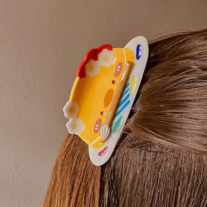 May include: A yellow and white hair clip in the shape of a painter's palette. The clip features a cartoon face, flowers, and a paintbrush. The palette has red, blue, and green paint. The clip is attached to brown hair.