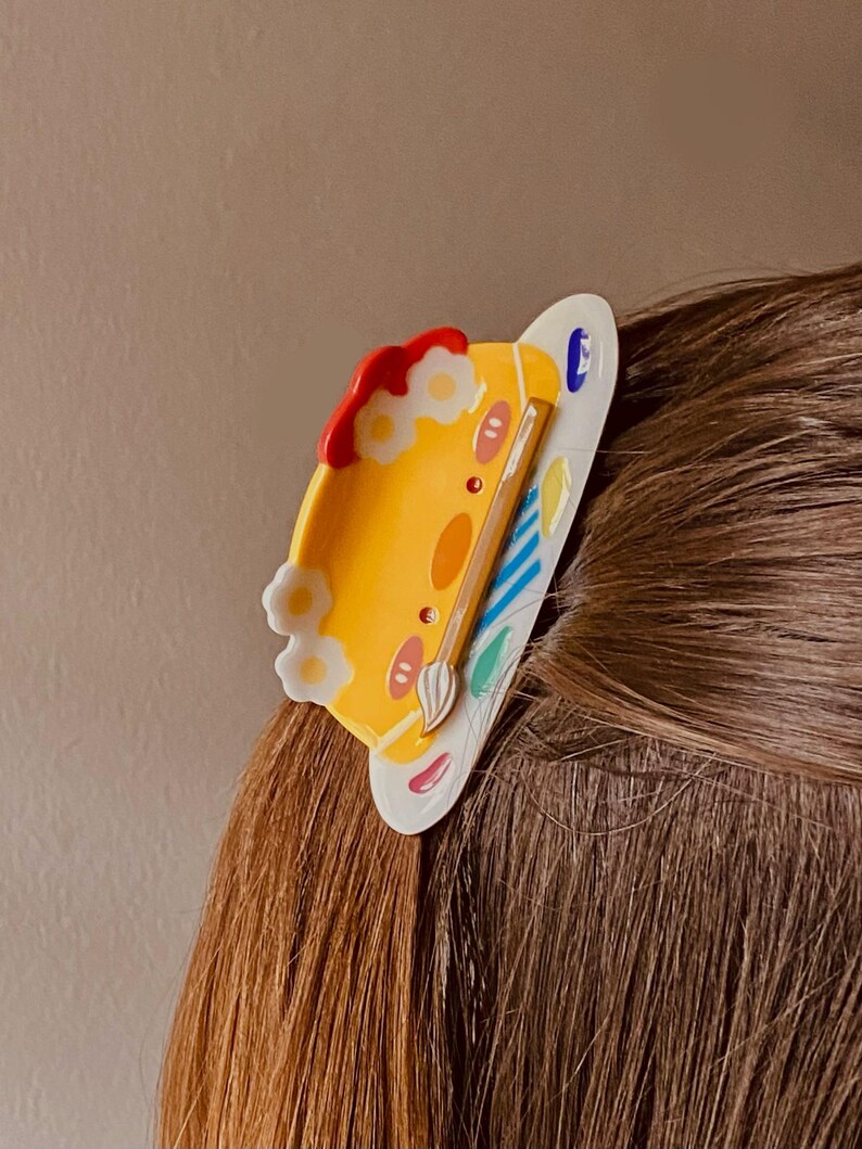 May include: A yellow and white hair clip in the shape of a painter's palette. The clip features a cartoon face, flowers, and a paintbrush. The palette has red, blue, and green paint. The clip is attached to brown hair.