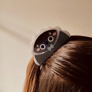 May include: A black and white hair clip with a cartoon face design. The clip has a scalloped edge and is secured in brown hair. The cartoon face has pink cheeks, blue eyes, and a star accent.