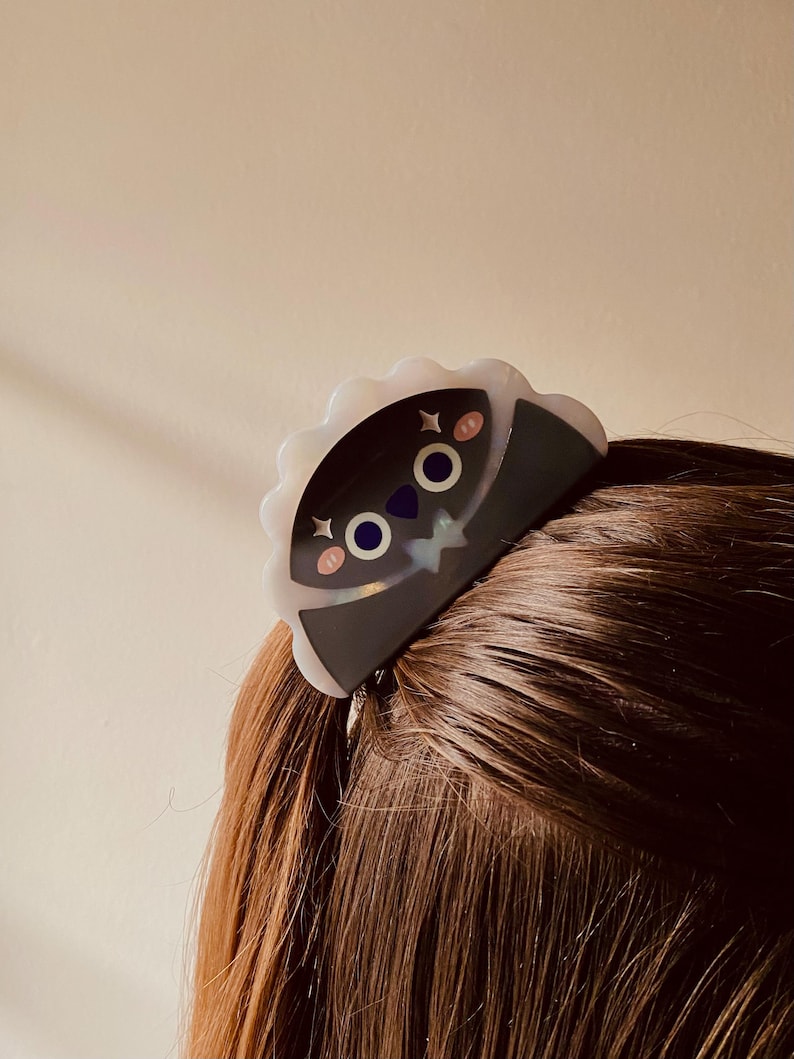 May include: A black and white hair clip with a cartoon face design. The clip has a scalloped edge and is secured in brown hair. The cartoon face has pink cheeks, blue eyes, and a star accent.