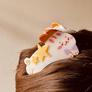 May include: A white and orange hair clip shaped like a cartoon cat with two yellow stars. The cat has pink cheeks and a purple bow. The clip is attached to brown hair.
