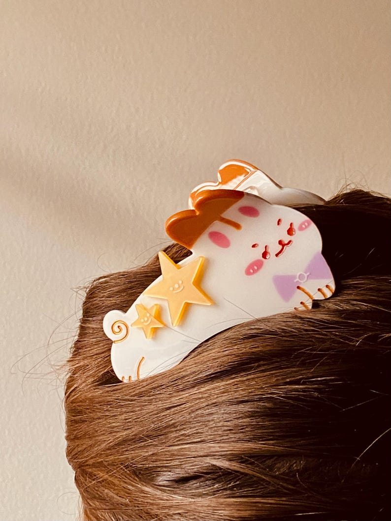 May include: A white and orange hair clip shaped like a cartoon cat with two yellow stars. The cat has pink cheeks and a purple bow. The clip is attached to brown hair.