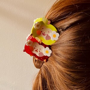 May include: Two colorful hair clips, one yellow and one red, each featuring a cartoon character design with white flower accents. The clips are set against a backdrop of brown hair.