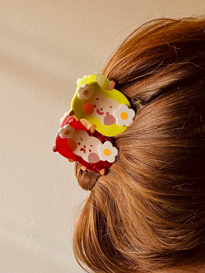 May include: Two colorful hair clips, one yellow and one red, each featuring a cartoon character design with white flower accents. The clips are set against a backdrop of brown hair.