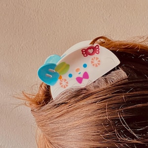 May include: A white hair clip with a cartoon design featuring a blue bow, a green and yellow leaf, and pink bow accents. The clip is decorated with small pink hearts and orange starbursts. The clip is placed in brown hair.