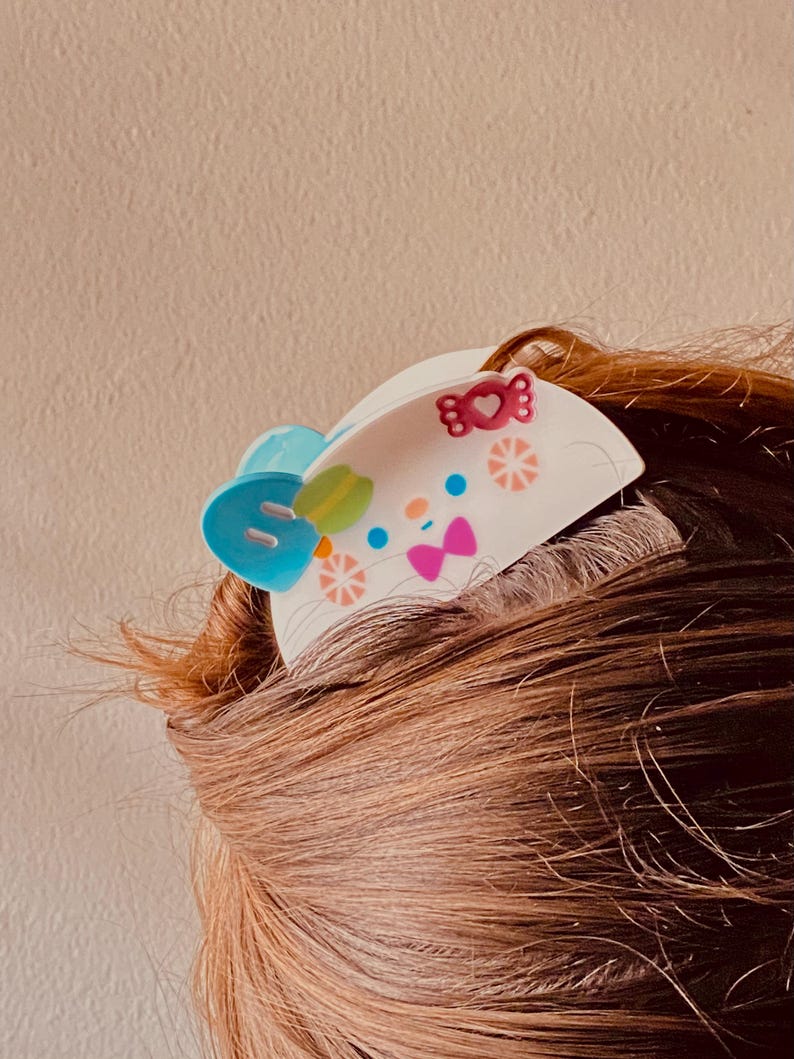 May include: A white hair clip with a cartoon design featuring a blue bow, a green and yellow leaf, and pink bow accents. The clip is decorated with small pink hearts and orange starbursts. The clip is placed in brown hair.